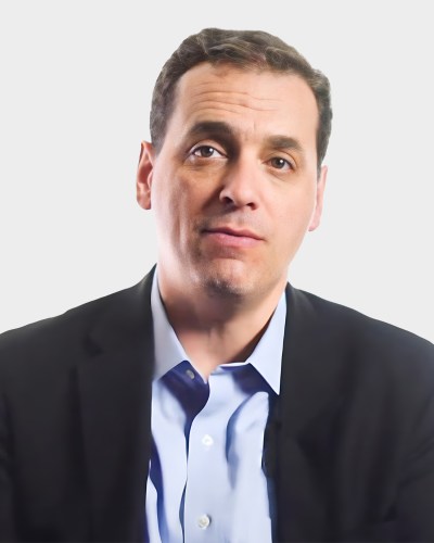 A man in a black suit and light blue shirt appears against a plain white background.