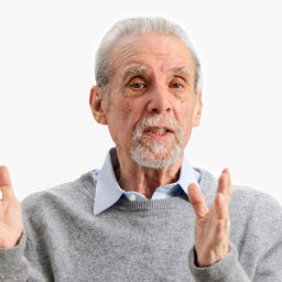 An elderly man gestures with his hands while speaking, wearing a gray sweater and blue shirt. The background is plain white with a logo in the top right corner.