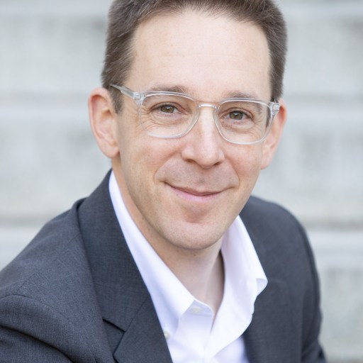 A person in a suit and glasses smiling, sitting on steps.