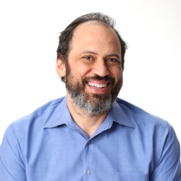 A bearded man with dark hair, wearing a blue shirt, smiles against a plain white background.