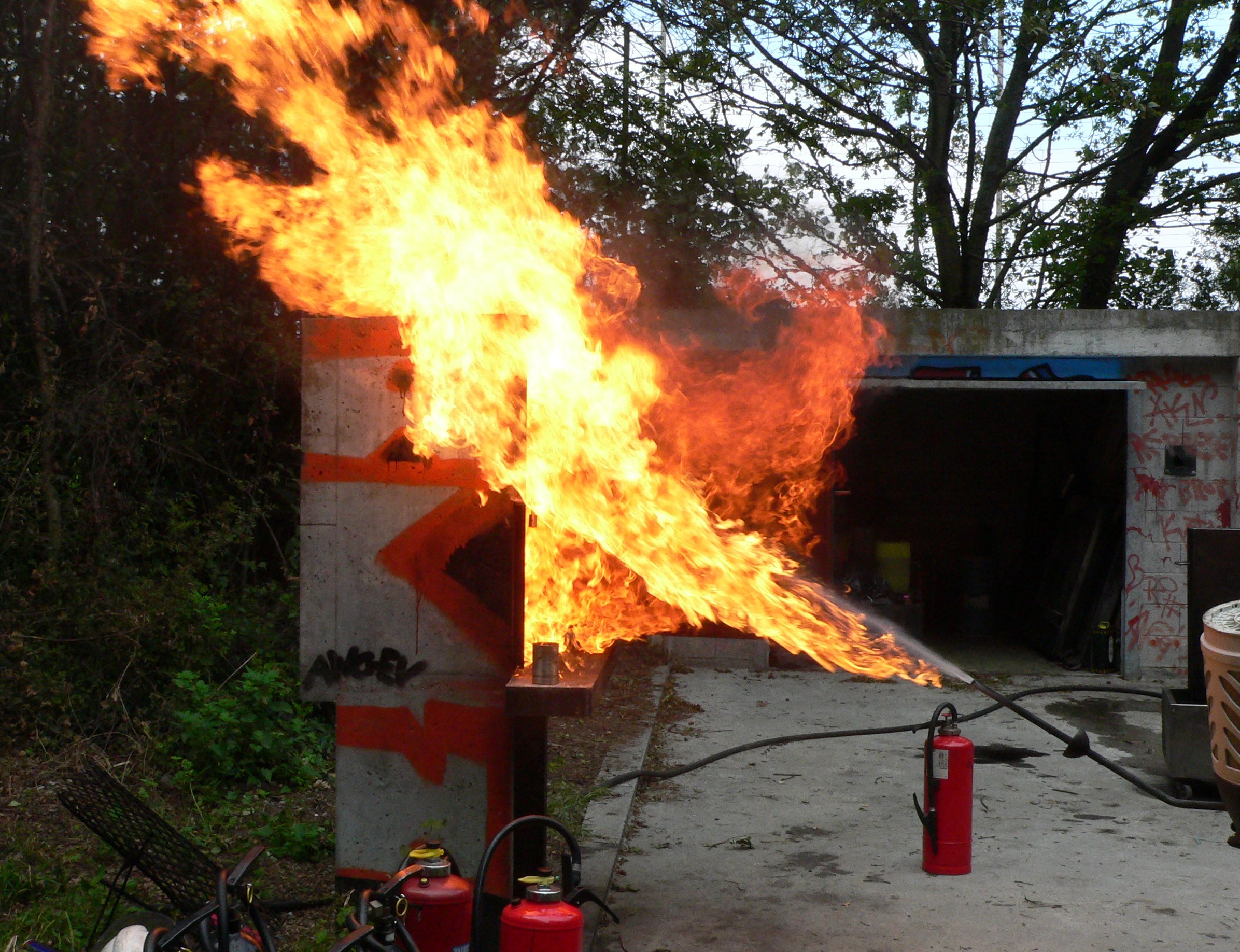 A large flame bursts from a structure outdoors, resembling an attempt to deep fry a frozen turkey gone wrong. Fire extinguishers are placed nearby, surrounded by trees and a graffitied wall. In the background, there's a garage standing witness to the chaotic scene.