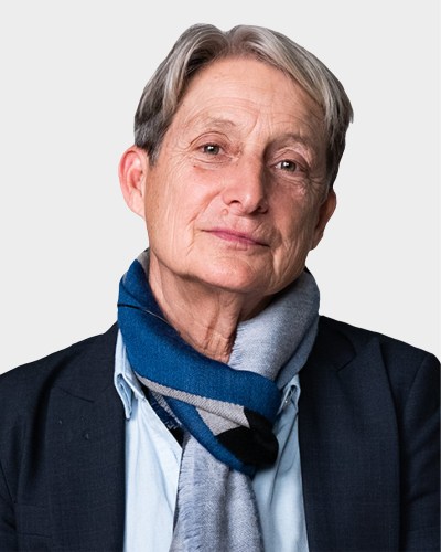 An individual with short hair wearing a suit and a blue scarf, looking at the camera with a neutral expression against a plain background.