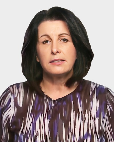 Person with shoulder-length dark hair wearing a patterned blouse against a plain background.