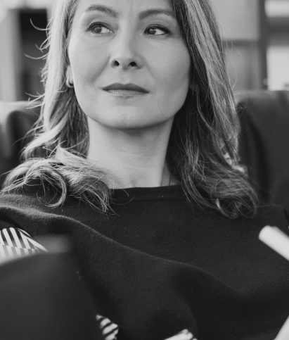 A black and white photo of a person with long hair sitting, looking to the side. They are wearing a dark top with a striped detail.