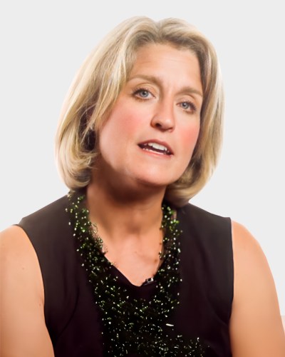 A person with short blonde hair, wearing a sleeveless black top and a green beaded necklace, is speaking against a plain background.