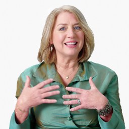 A person with shoulder-length blonde hair smiles, wearing a green blouse and gesturing with both hands.