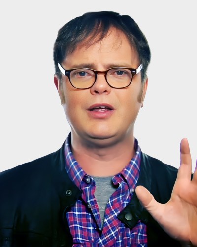 A person wearing glasses, a checkered shirt, and a jacket gestures with one hand raised.