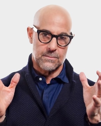 A person with glasses and a bald head, wearing a dark jacket and blue shirt, gestures with both hands against a plain background.