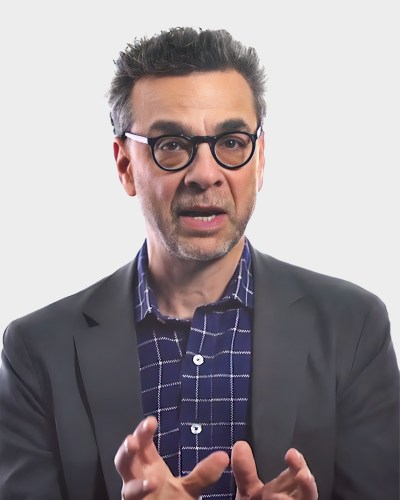 A person with glasses and a beard in a suit jacket and checkered shirt gestures with their hands against a plain background.