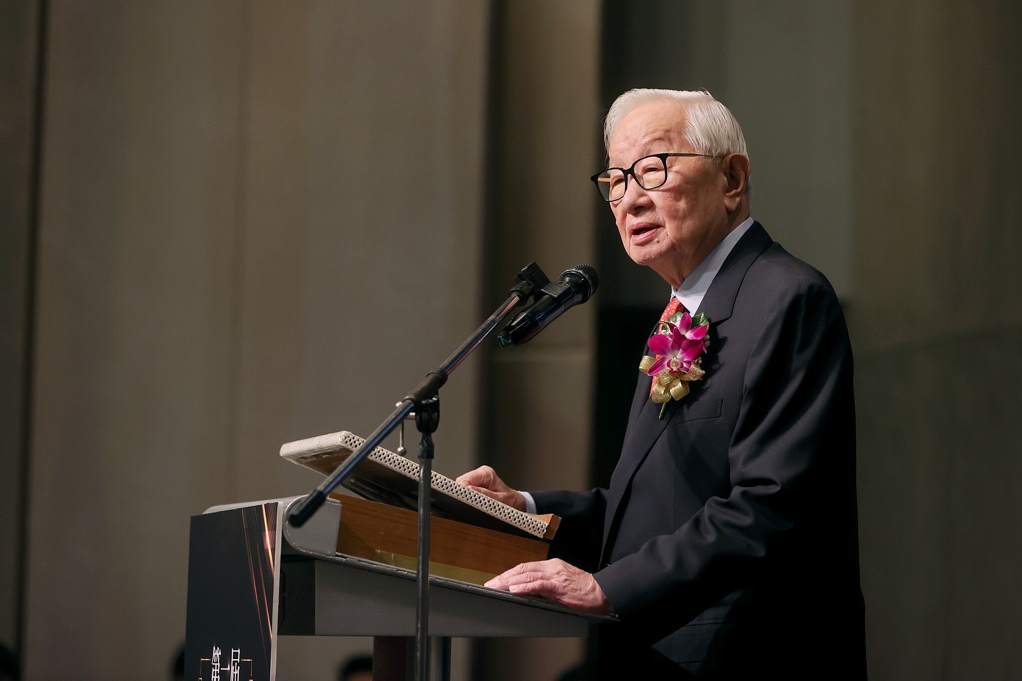 An elderly man in a suit and glasses stands at a podium with a microphone, speaking. He wears a red and purple flower corsage.