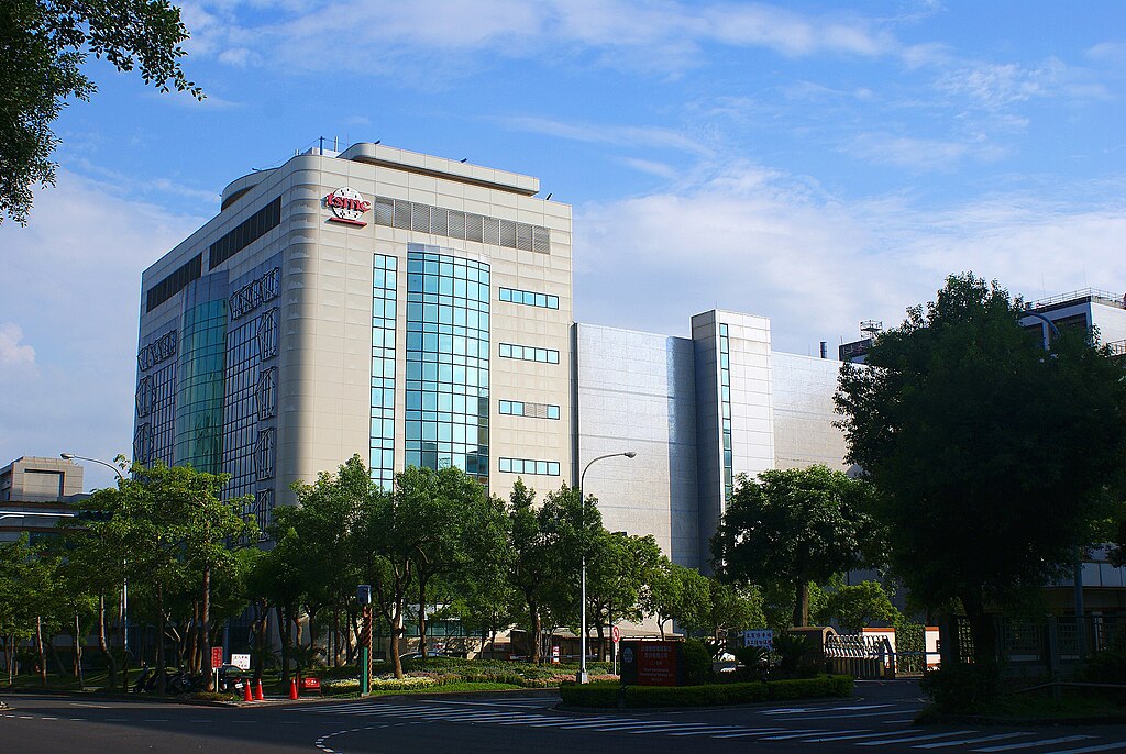 Modern office building with reflective glass and beige facade, surrounded by trees under a blue sky.