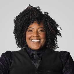 A person with curly hair smiles, wearing a dark vest and patterned shirt, against a plain background.