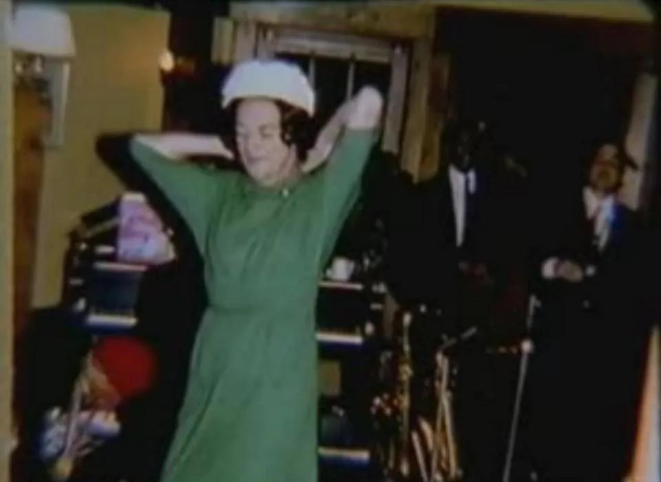 A woman in a green dress and white hat stands with arms raised like a conductor, evoking the energy of a musical hero, in a room with seated people and musical instruments in the background.