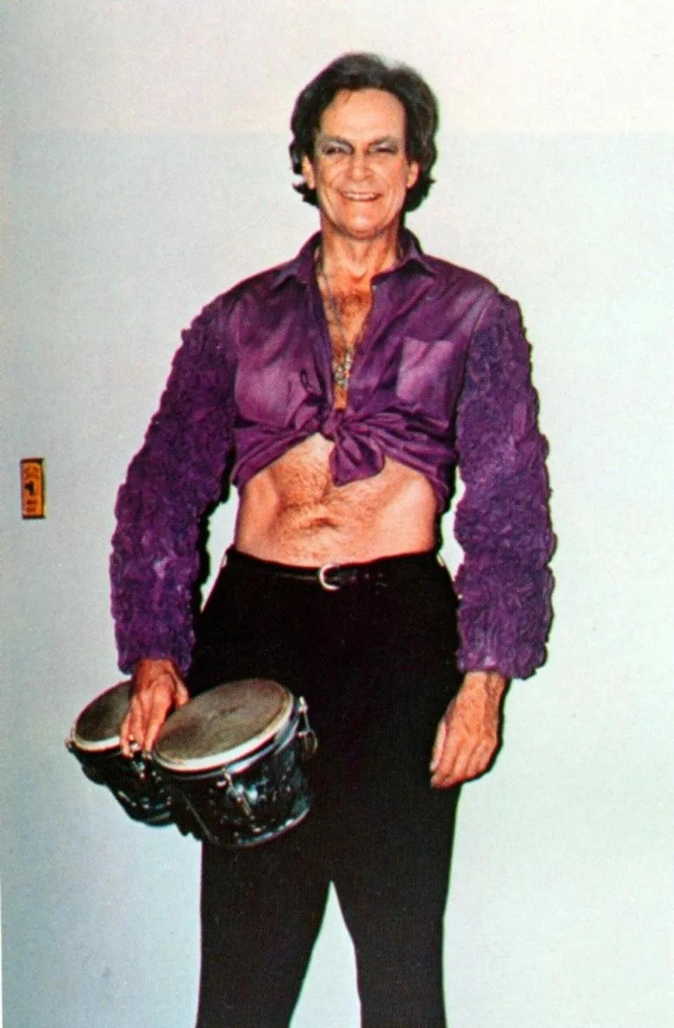 A person in a purple shirt tied at the front and black pants channels their inner hero as they hold a pair of bongo drums, smiling at the camera like Richard Feynman would.