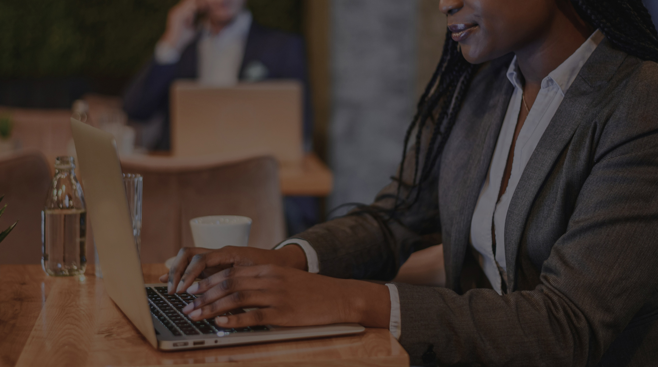 A person in a suit types on a laptop at a wooden table in a cafe. Another person in the background talks on a phone.