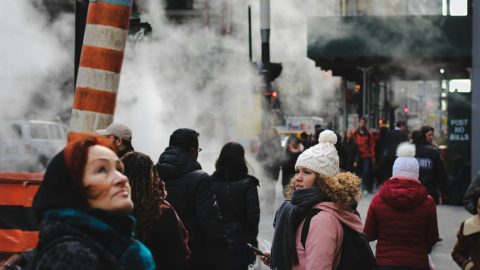 People walking on a city street with steam rising from vents create a scene reminiscent of an omics exposome research study. A woman in a white beanie looks back as buildings and traffic form the vibrant backdrop.