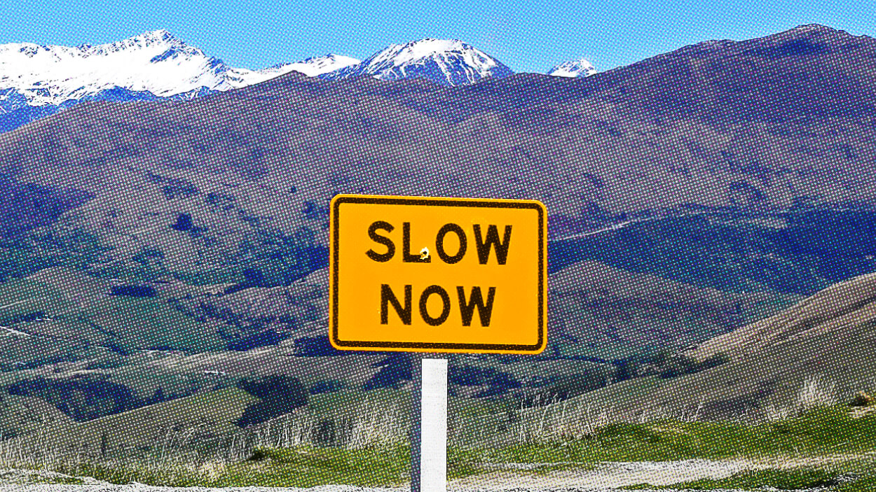 A yellow road sign reading "SLOW NOW" stands before a breathtaking mountainous landscape under a clear blue sky, reminding travelers of the art of slow productivity.