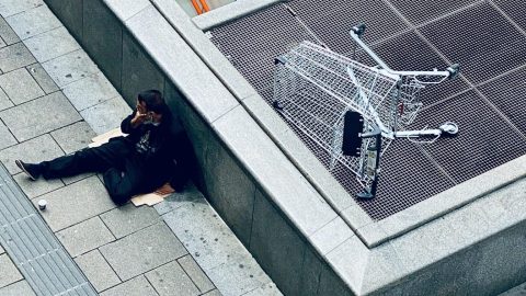 A person sits on a sidewalk near an escalator entrance, next to an overturned shopping cart on a metal grate.