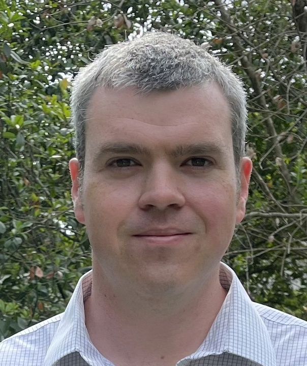 A person with short gray hair stands outdoors, wearing a patterned collared shirt. Green foliage is visible in the background.