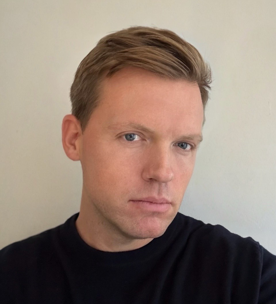 Portrait of a person with short light brown hair, wearing a black shirt, looking towards the camera with a neutral expression.