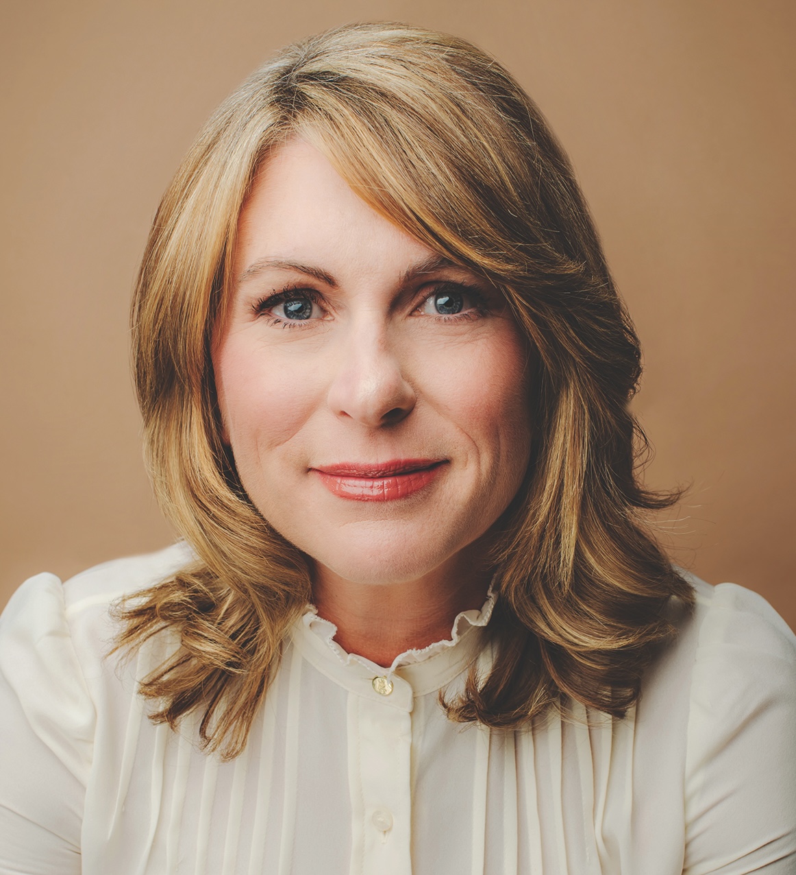 A person with shoulder-length blonde hair and a white blouse smiles slightly against a neutral background.