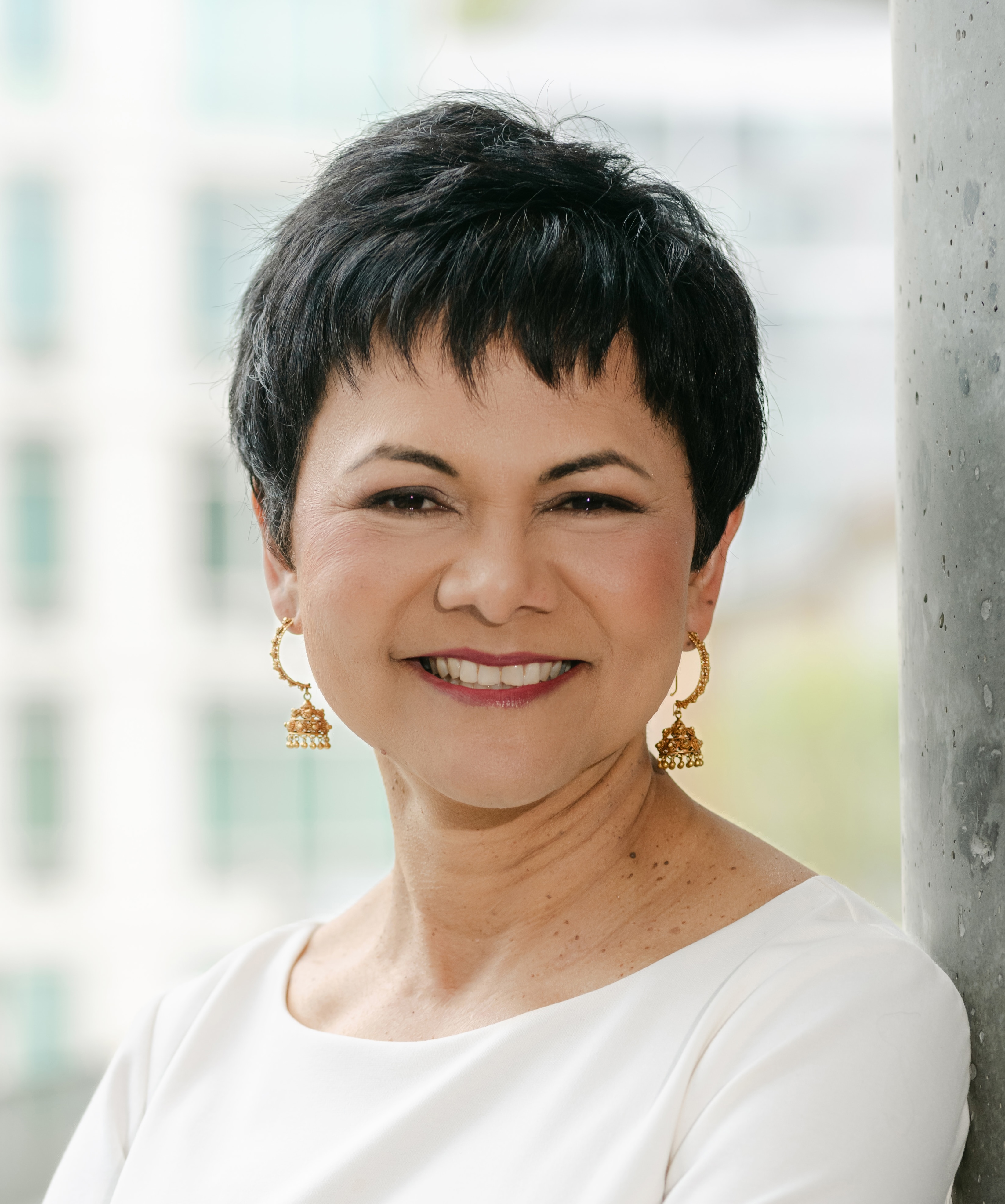 A woman with short black hair and gold earrings smiles, standing against a concrete pillar. She is wearing a white top. A blurred building is in the background.