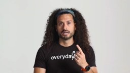 A person with long curly hair and a headband gestures with one hand, wearing a black shirt that says "everyday" and a smartwatch on the other wrist, against a plain background.