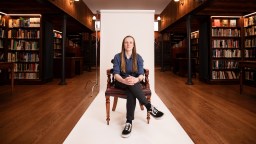 A person sits on a chair in a library setting, with bookshelves on either side and a white backdrop behind them.