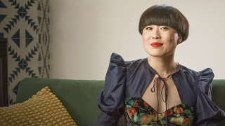 A woman with a bowl haircut and red lipstick sits on a sofa wearing a dark, floral dress. A textured yellow pillow and patterned wall are visible in the background.