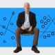A person sits on a stool against a blue background with black and white geometric diagrams, including lines, circles, squares, and triangles.