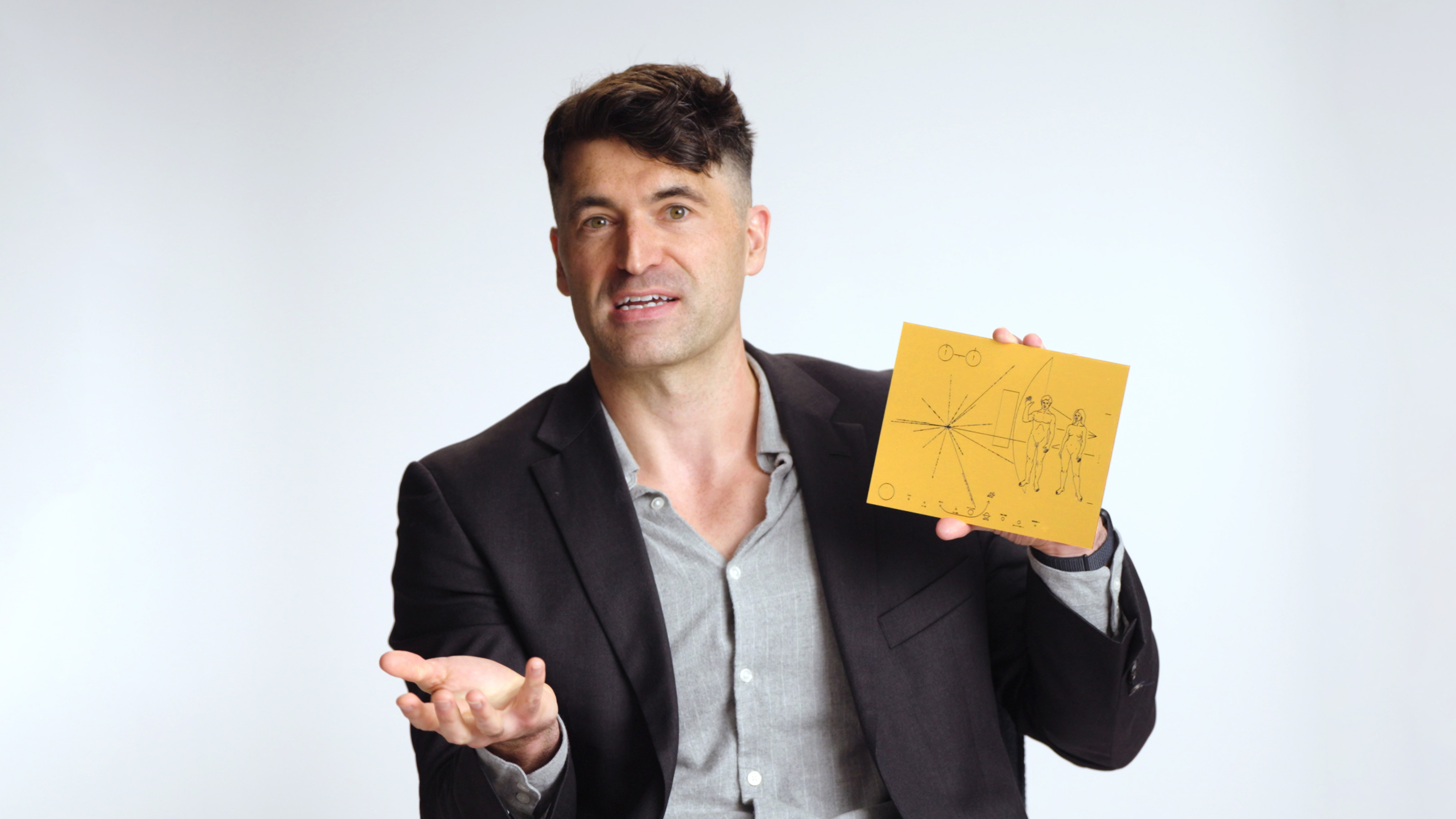 A man in a blazer holds up a yellow card with black line drawings and gestures with his other hand against a plain background.
