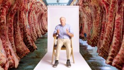 An older man sits on a chair in front of a white backdrop, surrounded by rows of hanging beef carcasses in a meat processing facility.
