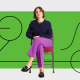A woman sits on a chair with legs crossed, wearing a navy sweater and purple pants, in front of a green background with black abstract line art.