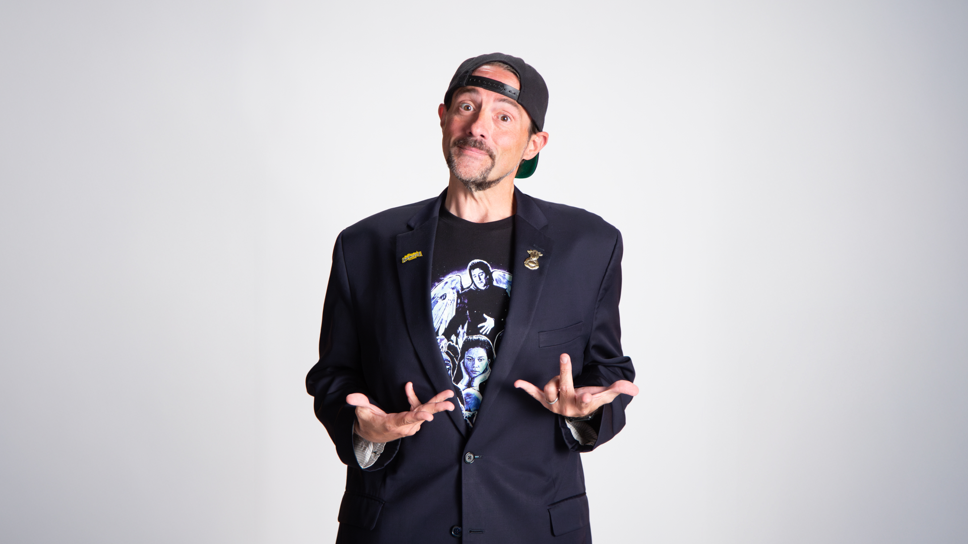 A man wearing a black blazer, graphic T-shirt, and backwards cap stands against a plain background with his hands raised in a questioning gesture.