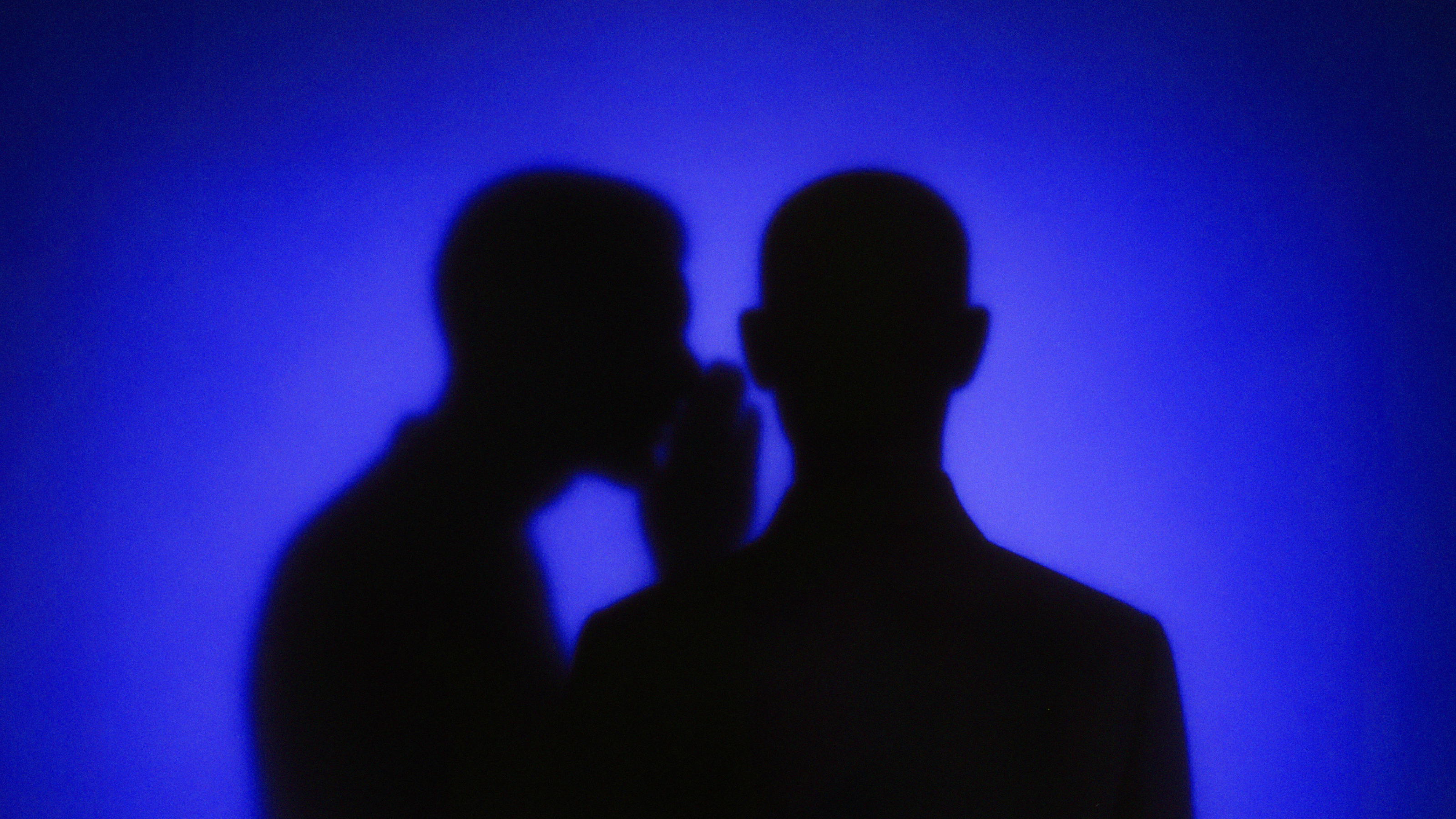 Two silhouetted figures against a blue background, with one appearing to whisper to the other.