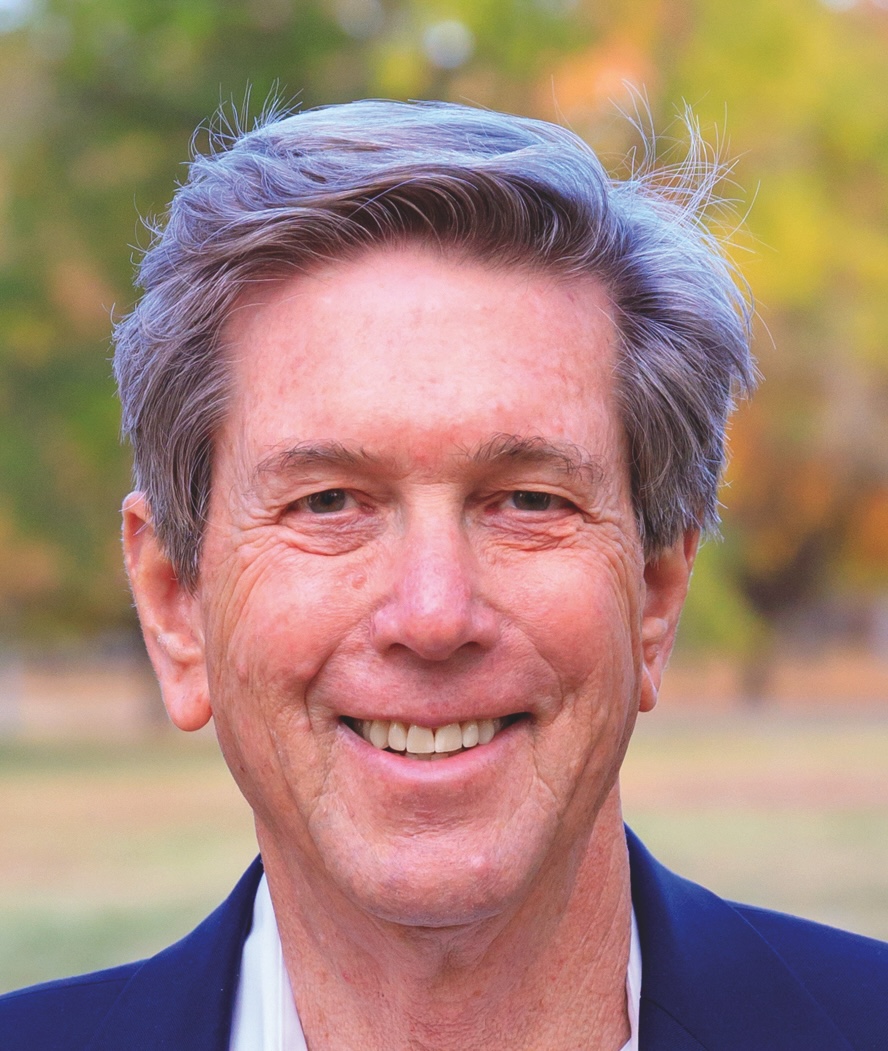 An older man with gray hair and a navy blazer smiles outdoors with blurred trees in the background.
