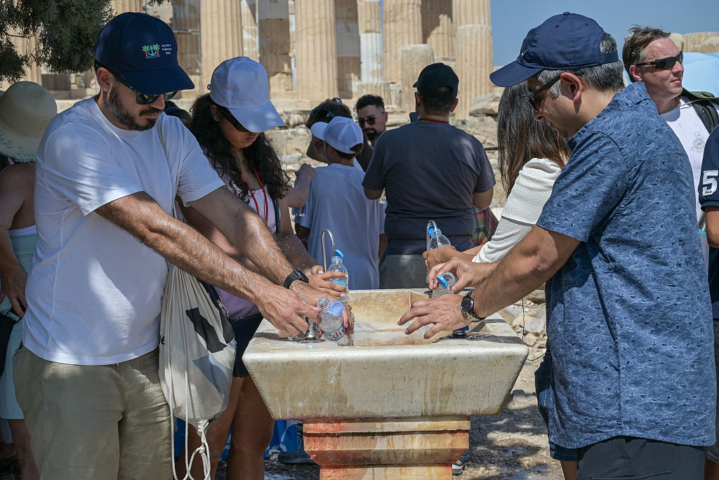Several people fill water bottles at a stone public fountain on a sunny day, with ancient columns in the background.