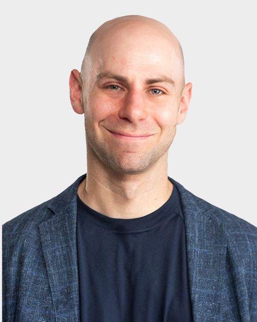 A bald man wearing a blue blazer and dark t-shirt smiles at the camera against a plain light background.
