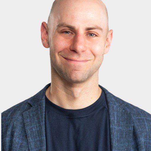A bald man wearing a blue blazer and dark t-shirt smiles at the camera against a plain light background.