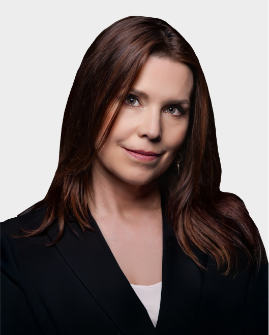 A woman with straight brown hair wearing a black blazer over a white top, posing against a plain, light background.