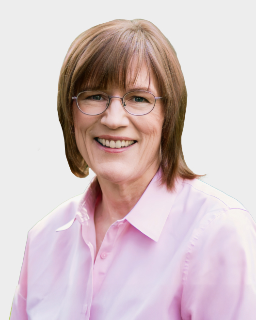 A person with short, light brown hair and glasses, wearing a light pink button-up shirt, smiling at the camera against a plain background.