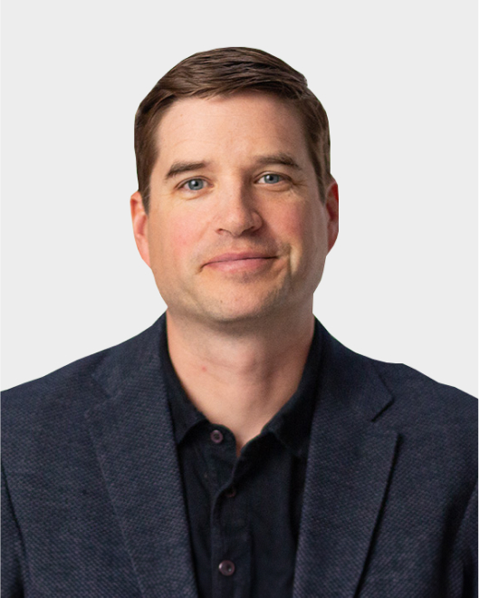 A man with short brown hair wearing a dark blazer and black shirt is facing the camera against a plain light background.
