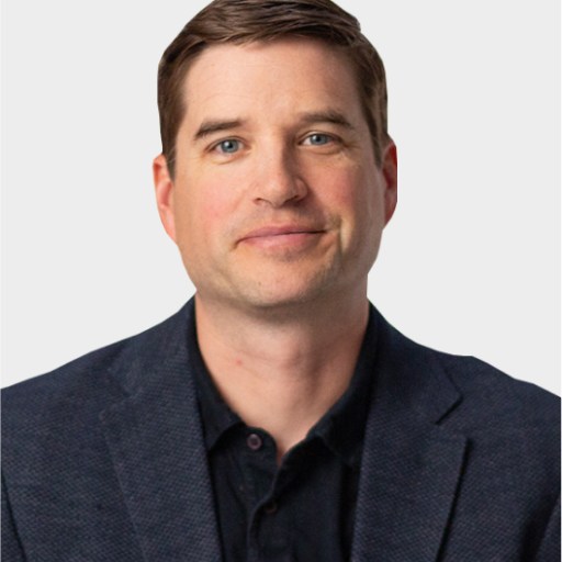 A man with short brown hair wearing a dark blazer and black shirt is facing the camera against a plain light background.