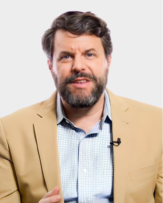 A man with a beard and mustache, wearing a beige blazer over a checkered shirt, sits in front of a plain white background with a lapel microphone clipped to his jacket.