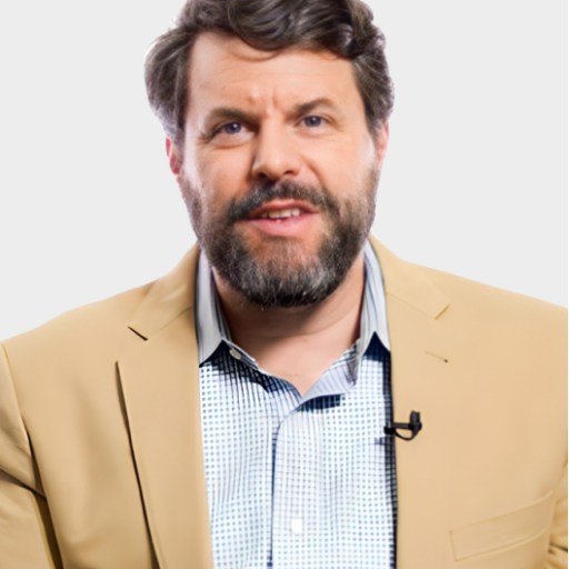 A man with a beard and mustache, wearing a beige blazer over a checkered shirt, sits in front of a plain white background with a lapel microphone clipped to his jacket.