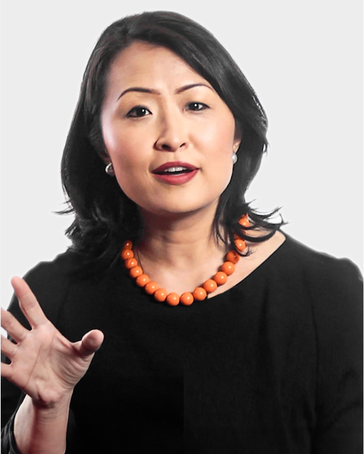 Woman with medium-length black hair wearing a black top and an orange beaded necklace, gesturing with her hand while speaking against a plain background.
