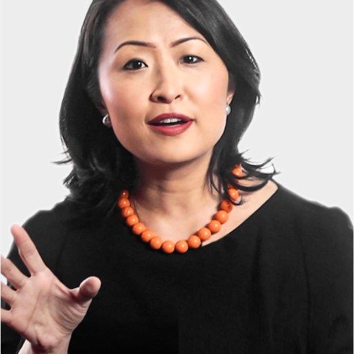 Woman with medium-length black hair wearing a black top and an orange beaded necklace, gesturing with her hand while speaking against a plain background.