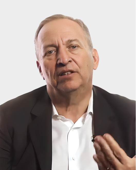 A middle-aged man in a suit jacket and white shirt speaks while gesturing with one hand against a plain, light background.