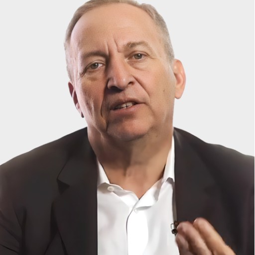 A middle-aged man in a suit jacket and white shirt speaks while gesturing with one hand against a plain, light background.
