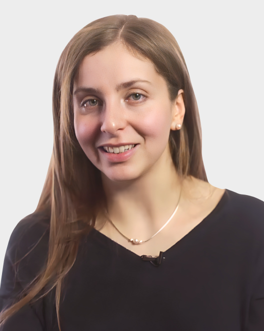 A woman with long brown hair wearing a black top and a necklace, smiling at the camera against a plain light background.